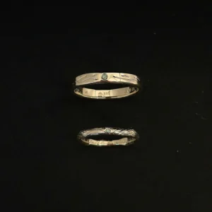 Zwei übereinander angeordnete, handgefertigte Goldringe. Der obere mir eingravierter Blätterranke, die von beiden Seiten auf einen grünen Brillanten zuläuft. Der Untere erinnert an einen Ast in dessen Astloch ein grüner Brillant eingefasst ist