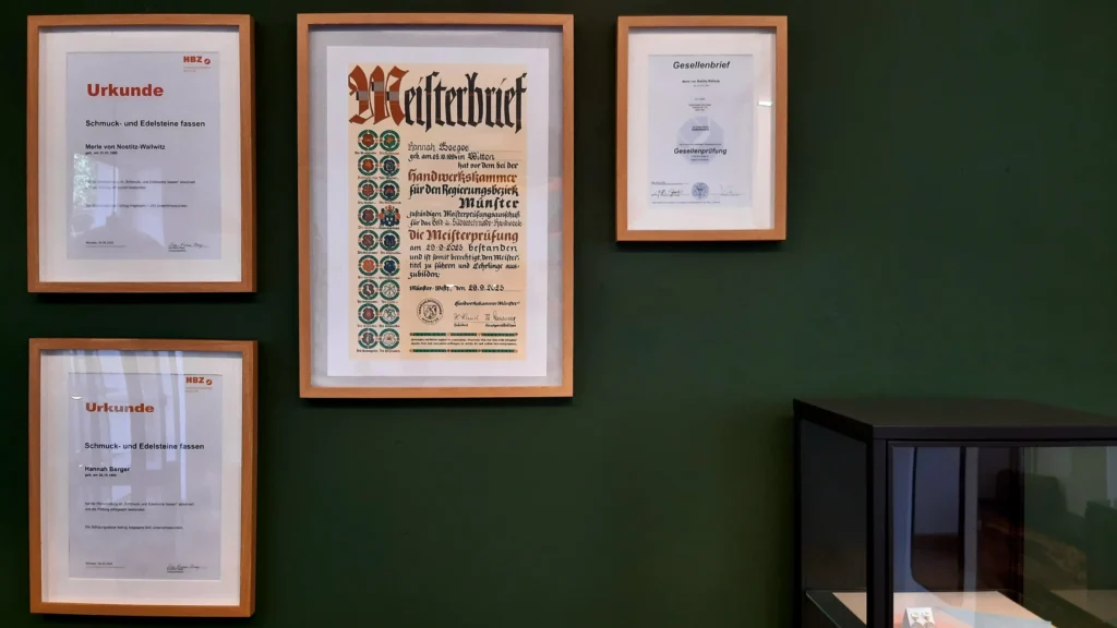 Vier Urkunden in Holzrahmen an einer dunkelgrünen Wand. Zwei Urkunden bescheinigen eine Schmuck- und Edelsteine fassen Weiterbildung, eine ist ein Meisterbrief der Gold- und Silberschmiede und die letzte ist ein Gesellenbrief der Gold- und Silberschmiede